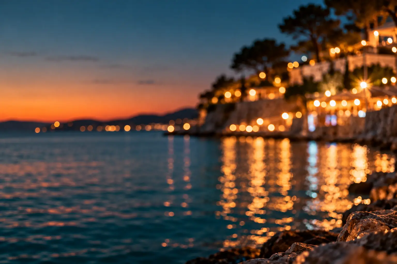 Unscharfe warme Lichter einer Küste spiegeln sich nachts im Meer, am Horizont letztes Abendrot