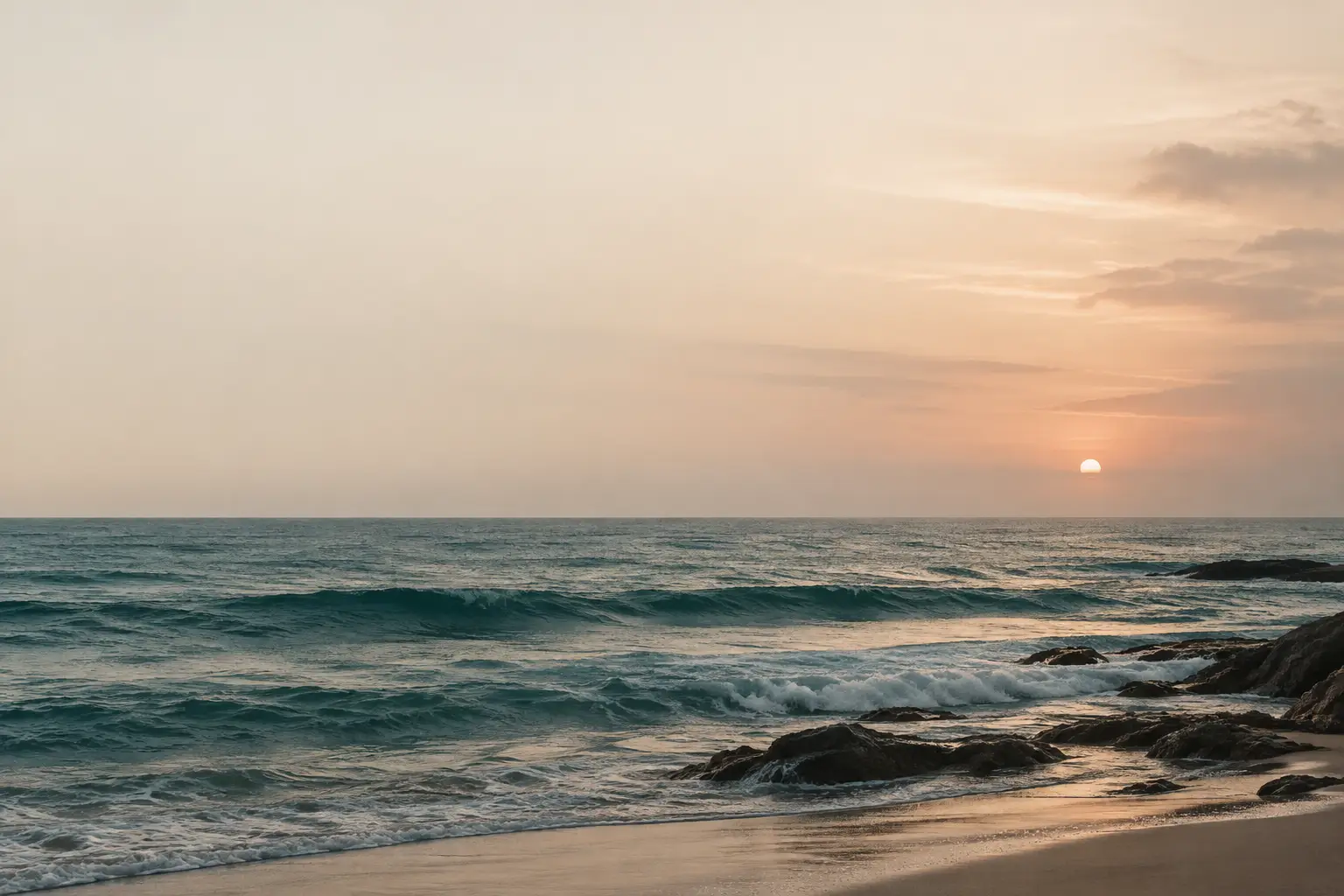 Blasser Sonnenuntergang über ruhigem Meer, sanfte Wellen, dunkle Felsen und heller Sandstrand im Dunst