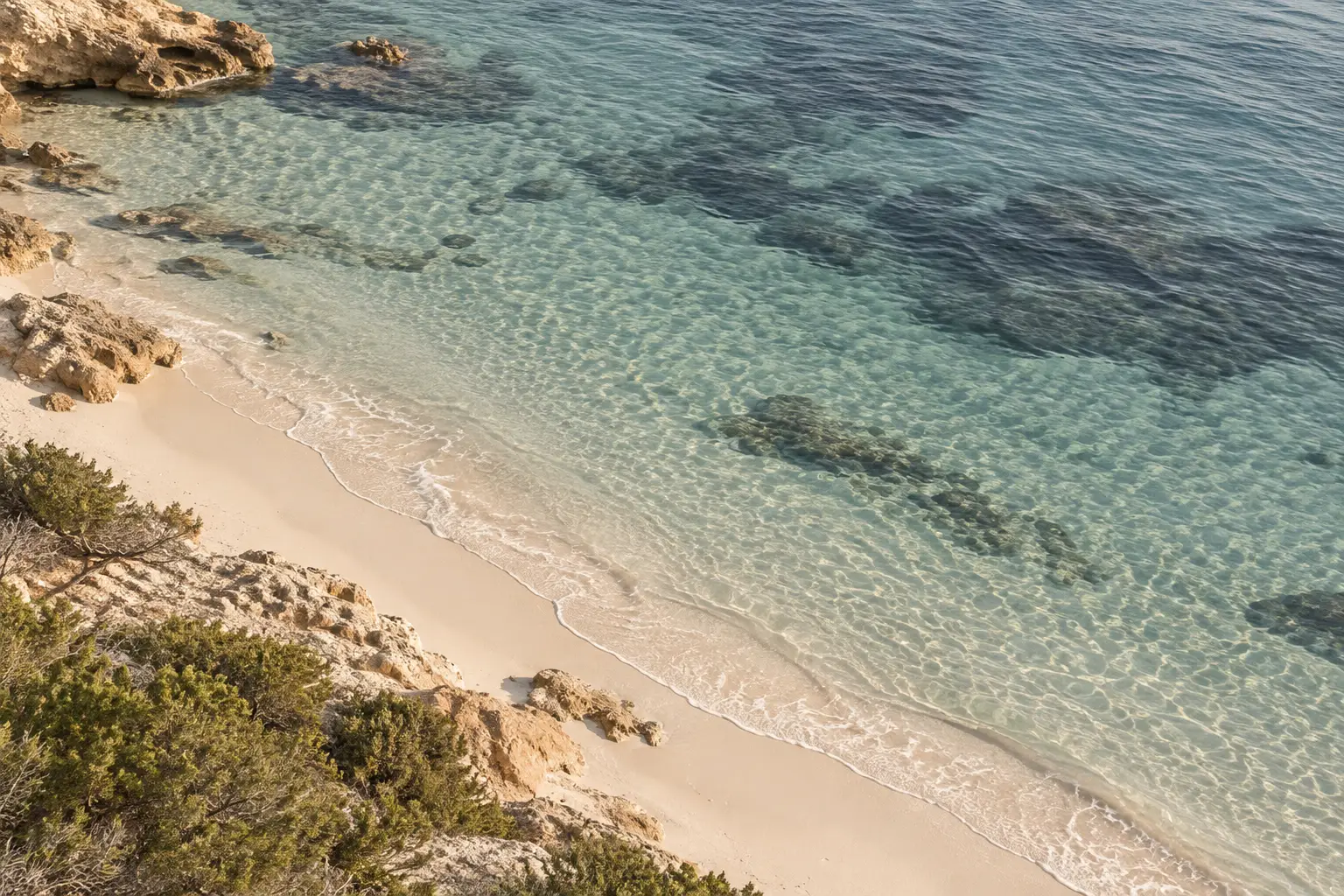 Blick von oben auf einen schmalen Sandstrand mit klarem, flachem Wasser, Felsen und Seegraswiesen am Grund