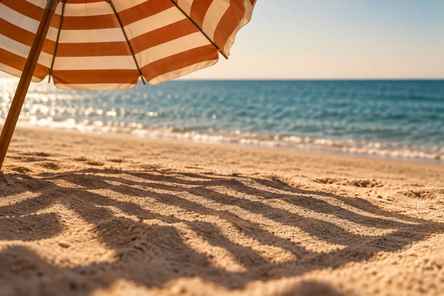Streifenschatten eines Sonnenschirms auf hellem Sand, dahinter unscharf das blaue Meer